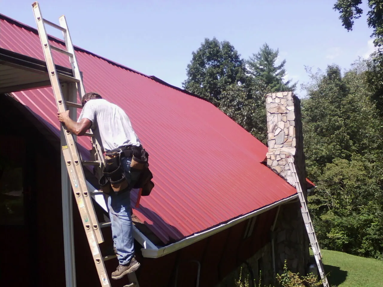 Roofer on a ladder beside a red metal roof and stone chimney — Soffit & Fascia Repair by Roofing Brothers in Fruitvale