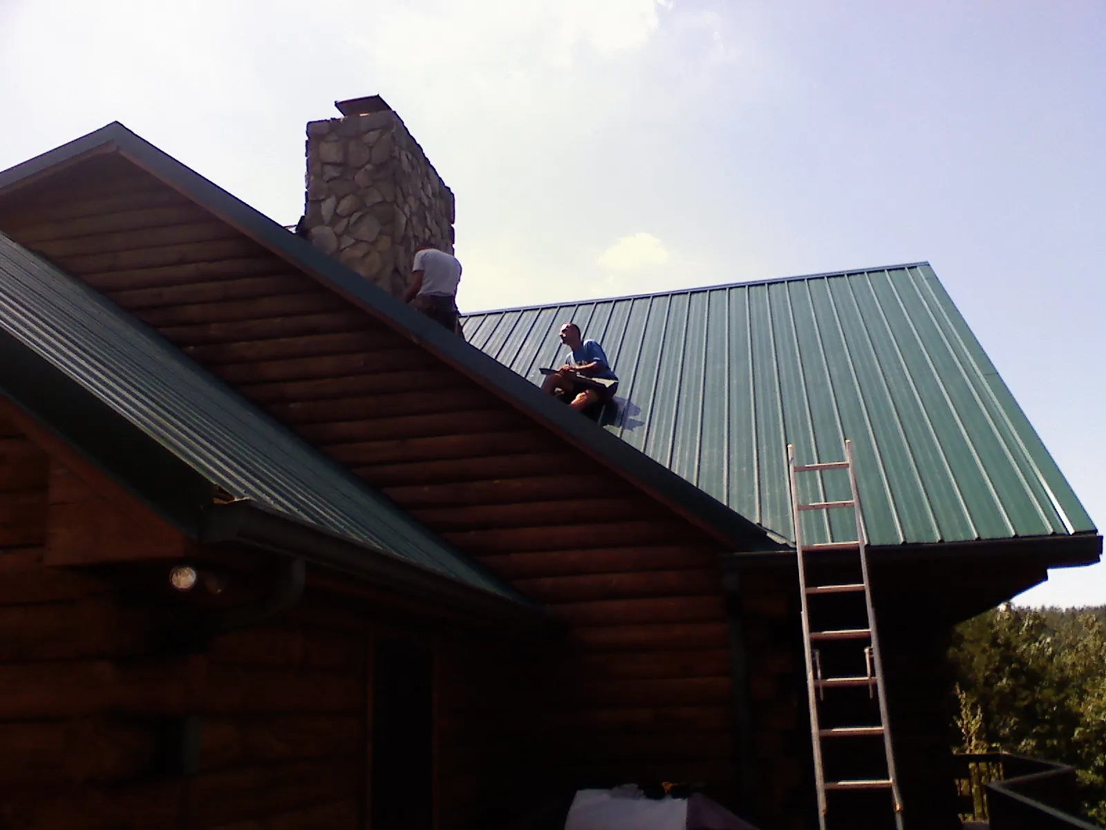 Roofers installing green metal panels beside a stone chimney on a log home in Fruitvale
